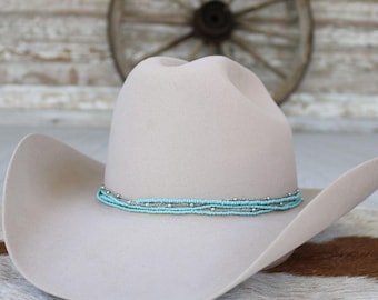 May include: A light beige cowboy hat with a turquoise and silver beaded band. The hat has a wide brim and a classic western design. The hat is likely made of felt or a similar material.