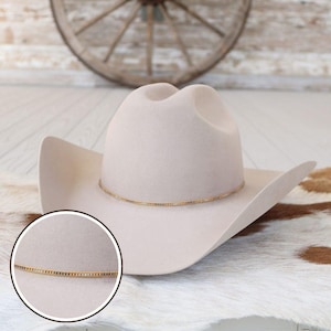 May include: A light beige cowboy hat with a gold-colored band around the crown. The hat has a classic western design with a wide brim and a rounded crown. The hat is displayed on a white and brown cowhide rug.