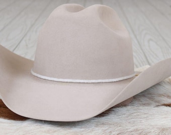 May include: A light beige felt cowboy hat featuring a high crown with a central crease and a wide, curved brim. A thin, white cord band encircles the base of the crown, providing a subtle contrast to the solid, neutral-toned felt material.