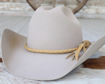 May include: A light beige cowboy hat with a braided leather band and decorative leather ties. The hat has a wide brim and a classic western design. The hat is made of a soft material.
