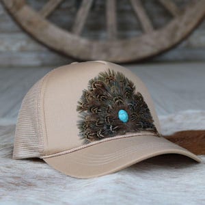 May include: A tan trucker hat with a brown and black pheasant feather design and a turquoise stone accent.