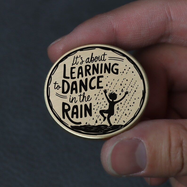Puede incluir: Un pin redondo de color dorado con texto negro y una silueta de una persona bailando bajo la lluvia. El texto dice "It's about learning to dance in the rain."