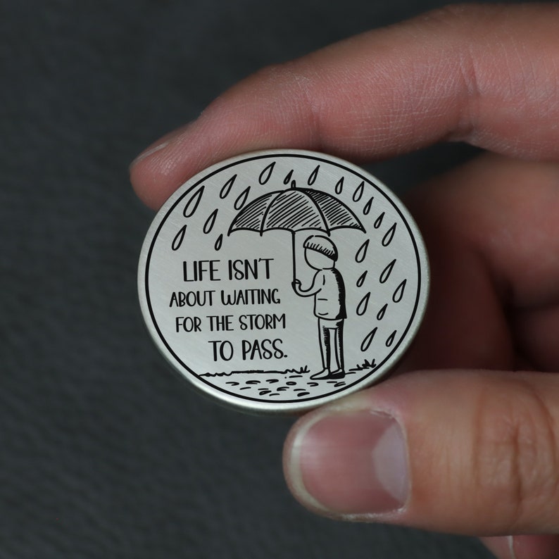 Puede incluir: Un token redondo plateado con una ilustraci&oacute;n en blanco y negro de una persona sosteniendo un paraguas bajo la lluvia. El texto en el token dice "Life isn't about waiting for the storm to pass."