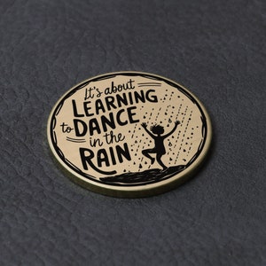 Puede incluir: Una moneda redonda de color dorado con una silueta negra de una persona bailando bajo la lluvia. El texto "It's about LEARNING to DANCE in the RAIN" est&aacute; impreso en la moneda.