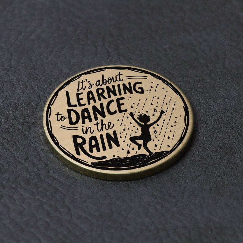 Puede incluir: Una moneda redonda de color dorado con una silueta negra de una persona bailando bajo la lluvia. El texto "It's about LEARNING to DANCE in the RAIN" est&aacute; impreso en la moneda.