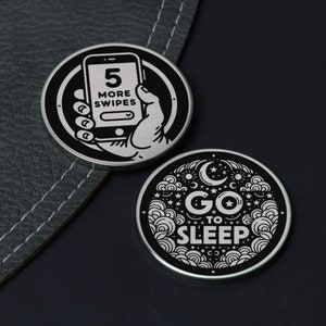 以下が含まれることがあります： 白のデザインが施された、黒い丸い金属製のトークンが2つ。最初のトークンは、スマートフォンを手に持つ手を描き、「5 more swipes」というテキストが書かれている。2番目のトークンは、三日月と星を描き、「Go to sleep」というテキストが書かれている。