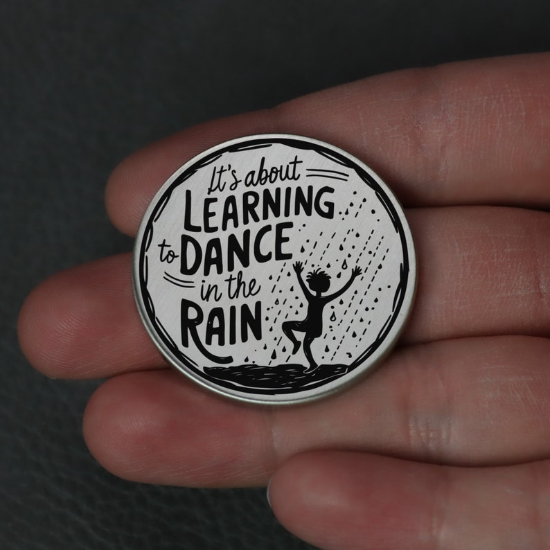 Puede incluir: Una moneda redonda de color plateado con una silueta negra de una persona bailando bajo la lluvia. La moneda tiene el texto "It's about learning to dance in the rain".