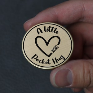 Puede incluir: Un token redondo de color dorado con el texto "A little Pocket Hug" y un corazón con "XOXO" en su interior.