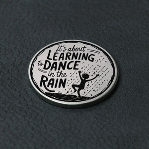 Puede incluir: Un pin redondo plateado con una silueta negra de una persona bailando bajo la lluvia. El texto en el pin dice "It's about learning to dance in the rain."