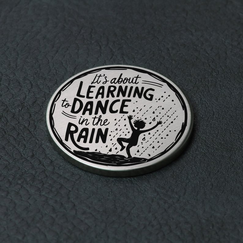 Puede incluir: Un pin redondo plateado con una silueta negra de una persona bailando bajo la lluvia. El texto en el pin dice "It's about learning to dance in the rain."