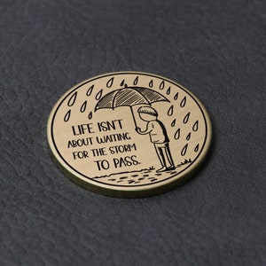 Puede incluir: Un token redondo de metal dorado con una ilustraci&oacute;n en blanco y negro de una persona sosteniendo un paraguas bajo la lluvia. El texto "LIFE ISN'T ABOUT WAITING FOR THE STORM TO PASS." est&aacute; grabado en el token.