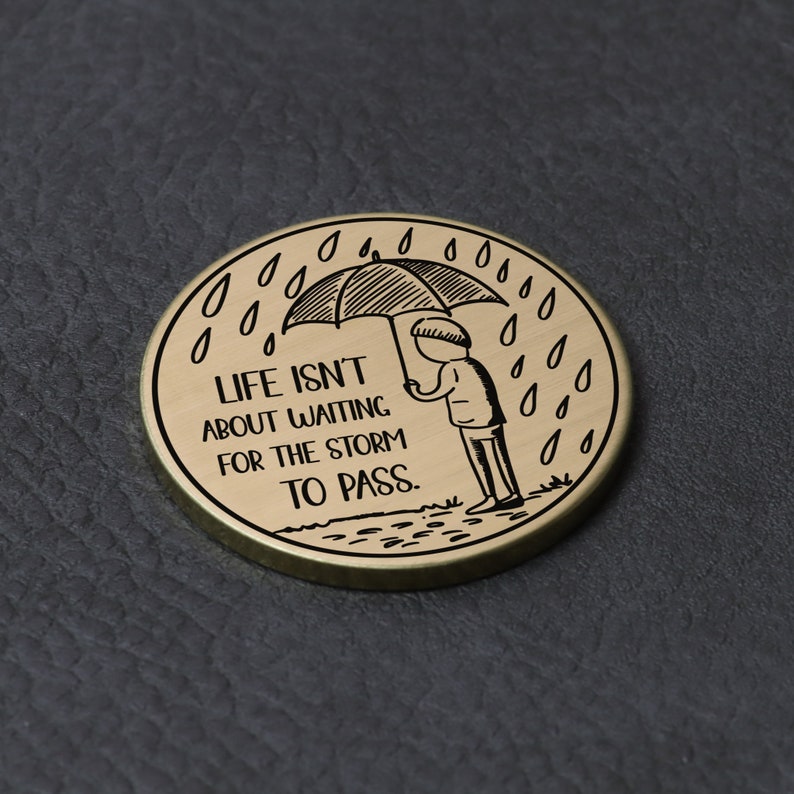 Puede incluir: Un token redondo de metal dorado con una ilustraci&oacute;n en blanco y negro de una persona sosteniendo un paraguas bajo la lluvia. El texto "LIFE ISN'T ABOUT WAITING FOR THE STORM TO PASS." est&aacute; grabado en el token.