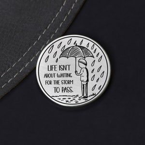 Puede incluir: Un token redondo de plata con una ilustraci&oacute;n en blanco y negro de una persona sosteniendo un paraguas bajo la lluvia. El texto en el token dice "Life isn't about waiting for the storm to pass."