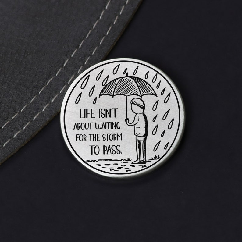 Puede incluir: Un token redondo de plata con una ilustraci&oacute;n en blanco y negro de una persona sosteniendo un paraguas bajo la lluvia. El texto en el token dice "Life isn't about waiting for the storm to pass."