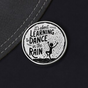Puede incluir: Un pin redondo plateado con una silueta negra de una persona bailando bajo la lluvia. El texto en el pin dice "It's about learning to dance in the rain."