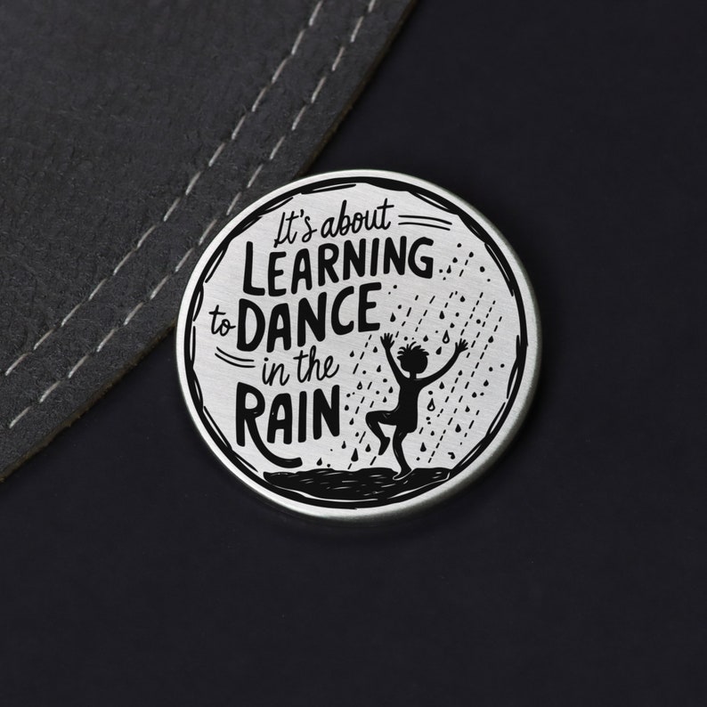 Puede incluir: Un pin redondo plateado con una silueta negra de una persona bailando bajo la lluvia. El texto en el pin dice "It's about learning to dance in the rain."
