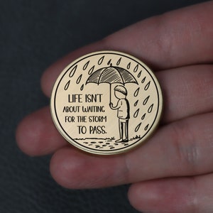 Puede incluir: Ficha redonda de color dorado con una ilustraci&oacute;n en blanco y negro de una persona sosteniendo un paraguas bajo la lluvia. El texto "LIFE ISN'T ABOUT WAITING FOR THE STORM TO PASS." est&aacute; inscrito en la ficha.