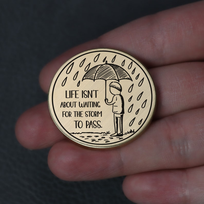 Puede incluir: Ficha redonda de color dorado con una ilustraci&oacute;n en blanco y negro de una persona sosteniendo un paraguas bajo la lluvia. El texto "LIFE ISN'T ABOUT WAITING FOR THE STORM TO PASS." est&aacute; inscrito en la ficha.