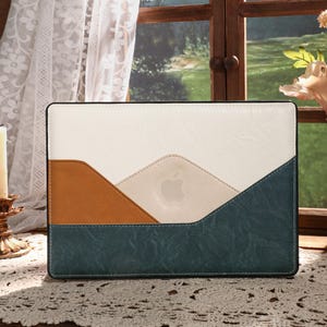 Puede incluir: Una funda rectangular para portátil con un diseño de bloques de color. La funda presenta una parte superior crema, una sección media marrón y una parte inferior verde azulado. El logotipo de Apple es visible en el centro. La funda está sobre un mantel de encaje.