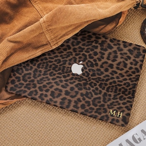 May include: A laptop with a leopard print design. The laptop has a white Apple logo and gold initials "M.H" on the bottom right corner. The laptop is partially covered by a brown corduroy jacket.