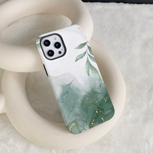 Puede incluir: Una funda de teléfono blanca con un diseño abstracto verde y dorado. El diseño presenta hojas y motas doradas sobre un fondo blanco.