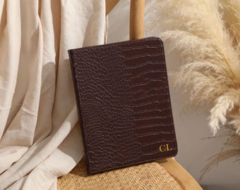Może przedstawiać: Ciemnobrązowe skórzane etui na tablet z teksturowanym wzorem przypominającym skórę krokodyla. Etui ma złoty monogram "CL" w prawym dolnym rogu. Etui spoczywa na plecionym krześle.