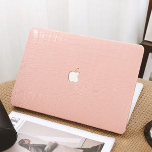 May include: A light pink laptop case with a crocodile skin texture. The case has a white Apple logo in the center. The laptop case is on a woven surface, with a black leather case and a magazine visible.