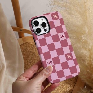 Puede incluir: Una funda de teléfono a cuadros rosa y granate con pequeños detalles de lazo. La funda se sostiene en una mano, mostrando el diseño. La funda del teléfono tiene un recorte para la cámara y está hecha de un material plástico duro.