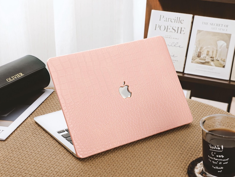 May include: A light pink laptop case with a crocodile skin texture. The case has a silver Apple logo in the center. The laptop is open, revealing the keyboard. The case is on a brown surface.