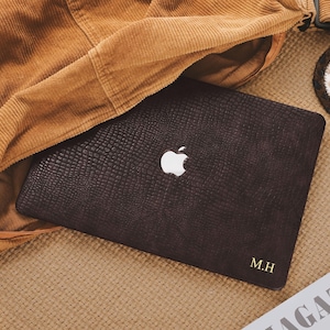 Könnte beinhalten: Eine dunkelbraune Laptop-Hülle mit einem strukturierten, reptilienartigen Muster. Das Apple-Logo ist sichtbar, zusammen mit den Initialen "M.H." in Gold. Die Hülle wird teilweise von einer beigen Cordjacke bedeckt.