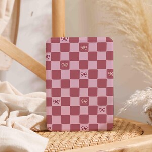 Puede incluir: Una funda de teléfono a cuadros rosa y burdeos con pequeñas ilustraciones de lazos. La funda tiene esquinas redondeadas y se muestra sobre una superficie tejida, lo que sugiere un artículo decorativo o de moda.