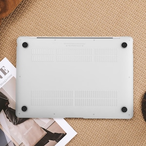 May include: A white laptop case with black rubber feet, resting on a textured beige surface. The case has ventilation slots and is surrounded by a magazine, a black bag, and a coaster with a cup of coffee.