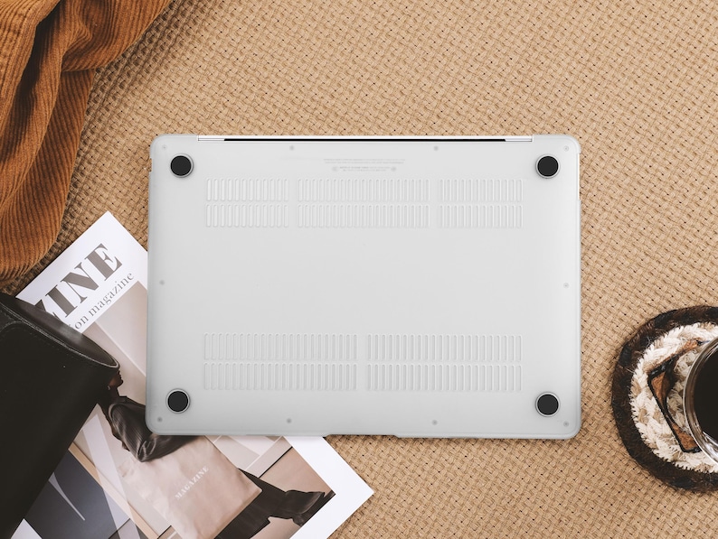 May include: A white laptop case with black rubber feet, resting on a textured beige surface. The case has ventilation slots and is surrounded by a magazine, a black bag, and a coaster with a cup of coffee.
