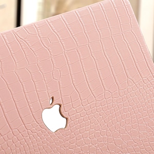 May include: A light pink laptop case with a crocodile skin texture. The case features a cutout for the Apple logo. The case is designed to protect the laptop from scratches and other damage.
