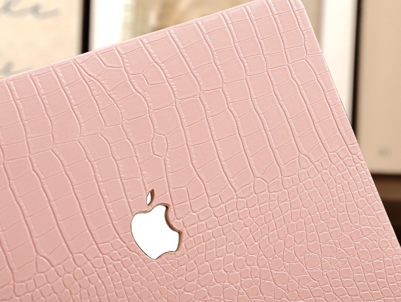 May include: A light pink laptop case with a crocodile skin texture. The case features a cutout for the Apple logo. The case is designed to protect the laptop from scratches and other damage.