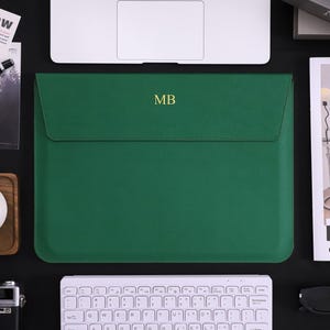 May include: A green laptop sleeve with a flap closure, featuring the gold initials "MB". The sleeve is surrounded by a white keyboard, a camera, a cup of coffee, and a magazine. The overall aesthetic is modern and minimalist.