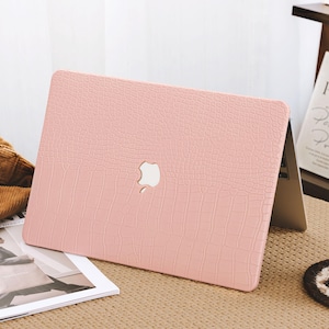 May include: A light pink laptop case with a textured, crocodile-like pattern. The case features a white Apple logo and is placed on a light brown surface. A magazine and a cup of coffee are also visible.