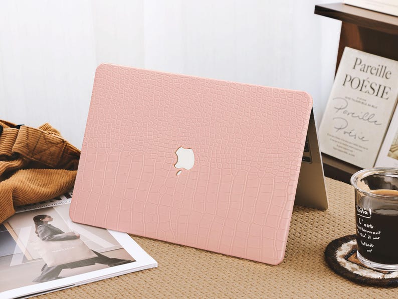 May include: A light pink laptop case with a textured, crocodile-like pattern. The case features a white Apple logo and is placed on a light brown surface. A magazine and a cup of coffee are also visible.