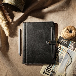 Puede incluir: Un diario de cuero negro con cierre dorado y un bolígrafo sobre una superficie de tela marrón.