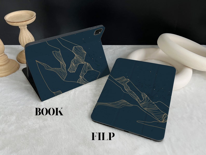 May include: Two dark blue phone cases with a gold line design. The design is of a mountain range. The text "BOOK" and "FILP" are printed on the white surface below the cases.