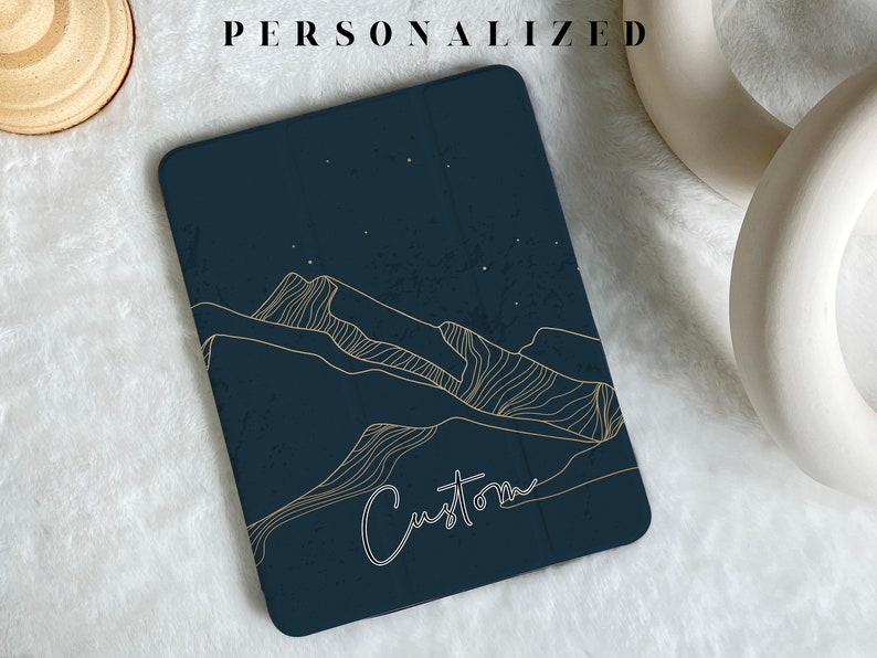 May include: A dark blue tablet case with a gold outline of mountains and stars. The text "Custom" is written in white cursive on the case.