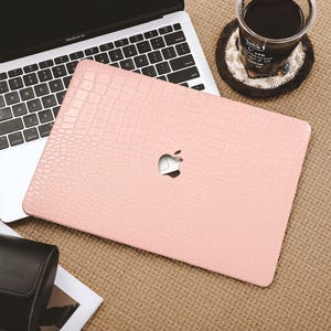 May include: A light pink laptop case with a crocodile skin texture, featuring a cutout for the Apple logo. The case is placed on top of a MacBook Air, with a cup of coffee and a black clutch bag nearby. The overall theme is tech accessories.