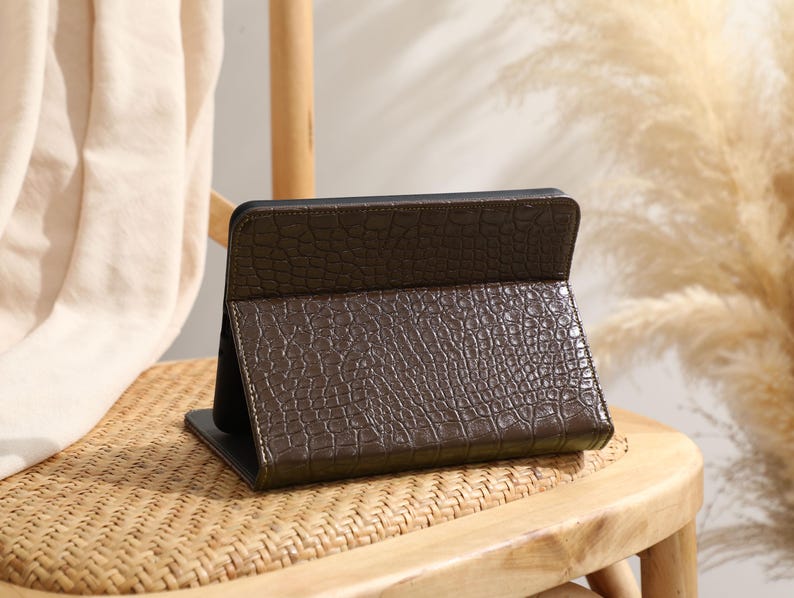 Może przedstawiać: Brązowe etui na tablet z teksturowanym wzorem przypominającym sk&oacute;rę krokodyla. Etui jest otwarte i oparte na drewnianym krześle. Etui jest w kolorze ciemnobrązowym z czarnym wnętrzem.