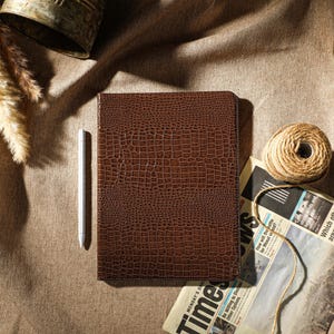 May include: A brown, crocodile-textured leather journal sits on a textured surface. A silver stylus lies next to it. A ball of twine and a newspaper are also present, suggesting a stationery theme.