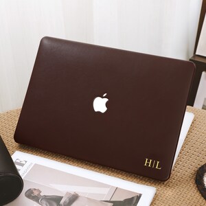 Op de afbeelding: Een bruine laptop met een wit Apple-logo en gouden initialen "H|L". De laptop staat op een beige oppervlak, vergezeld van een tijdschrift en een zwarte etui. Een kopje koffie staat op een onderzetter.