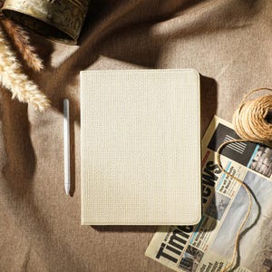 Op de afbeelding: Een witte, textuurrijke notebookhoes met een witte stylus pen op een bruine stoffen achtergrond. Een krant en een bol touw zijn ook zichtbaar op de achtergrond.
