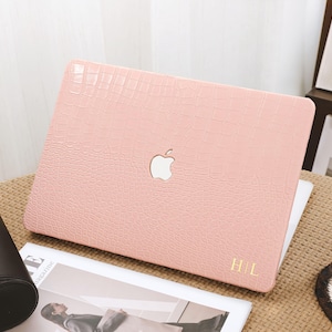 May include: A pink laptop case with a crocodile skin texture. The case has a cut-out for the Apple logo and gold initials "HL" in the corner. The laptop case is on a table with a magazine and a cup of coffee.