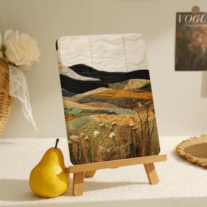 Peut inclure: Une peinture de paysage encadrée avec un motif texturé et abstrait de collines et de champs dans des tons de brun, de vert et de blanc. La peinture est sur un petit chevalet en bois.