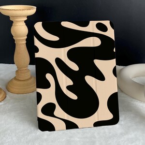 Puede incluir: Una funda para tableta con un fondo beige y un patrón abstracto negro. La funda está de pie sobre una superficie blanca. En el fondo se ven candelabros decorativos de madera.