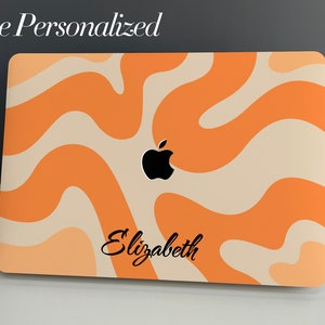 May include: A laptop computer with a personalised orange and cream abstract design. The Apple logo is visible on the lid, and the name "Elizabeth" is printed on the bottom of the laptop.